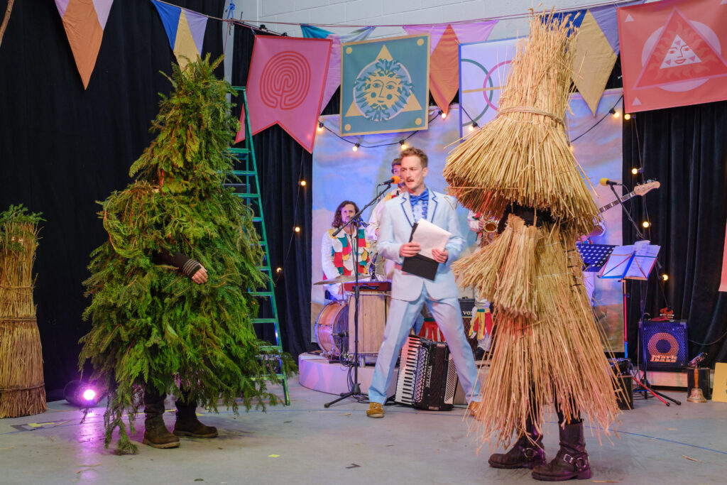 Two people dressed as a hedge and a bush with a man in a blue suit speaking into a microphone.